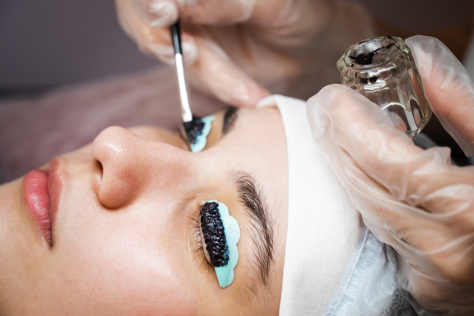 Close-up portrait of a woman on eyelash lamination procedure. The master applies tint to the eyelashes.